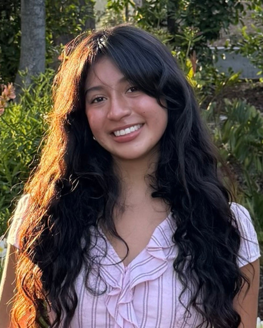 Girl standing in front of bushes wearing pink blouse with long brown hair.