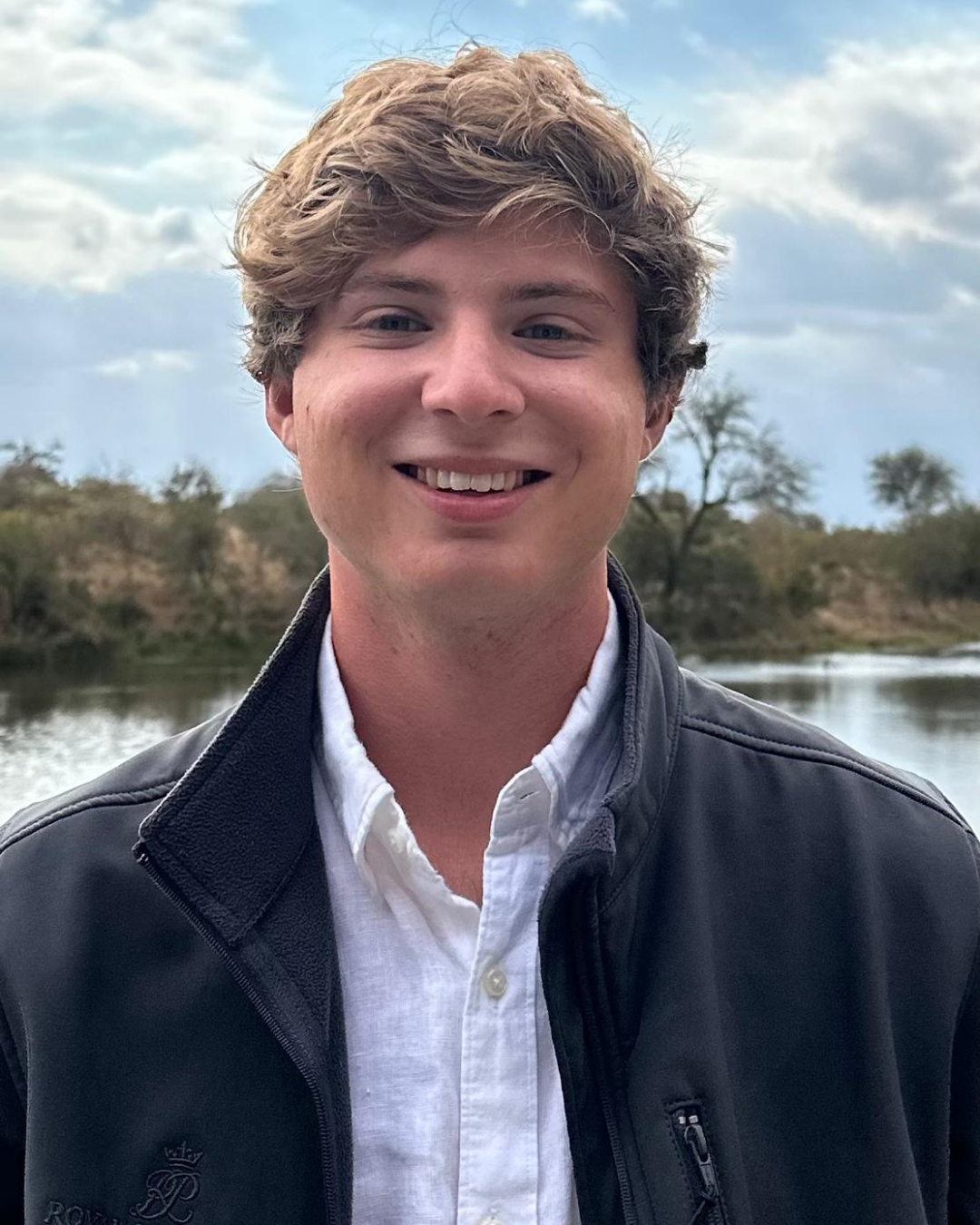 Man wearing navy jacket with short blonde curly hair standing in front of lake.