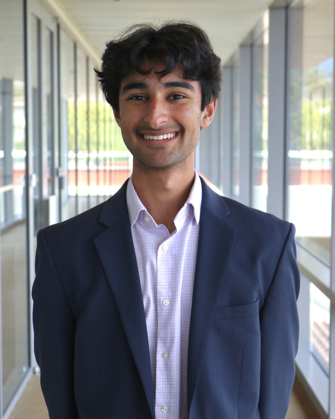 Man wearing navy blazer with short black curly hair standing in front of walkway. 