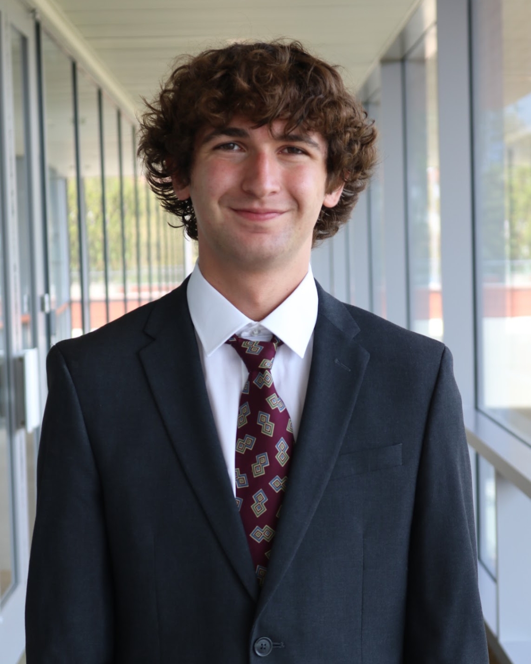 Man in black suit with red tie and brown curly hair standing in front of walkway.