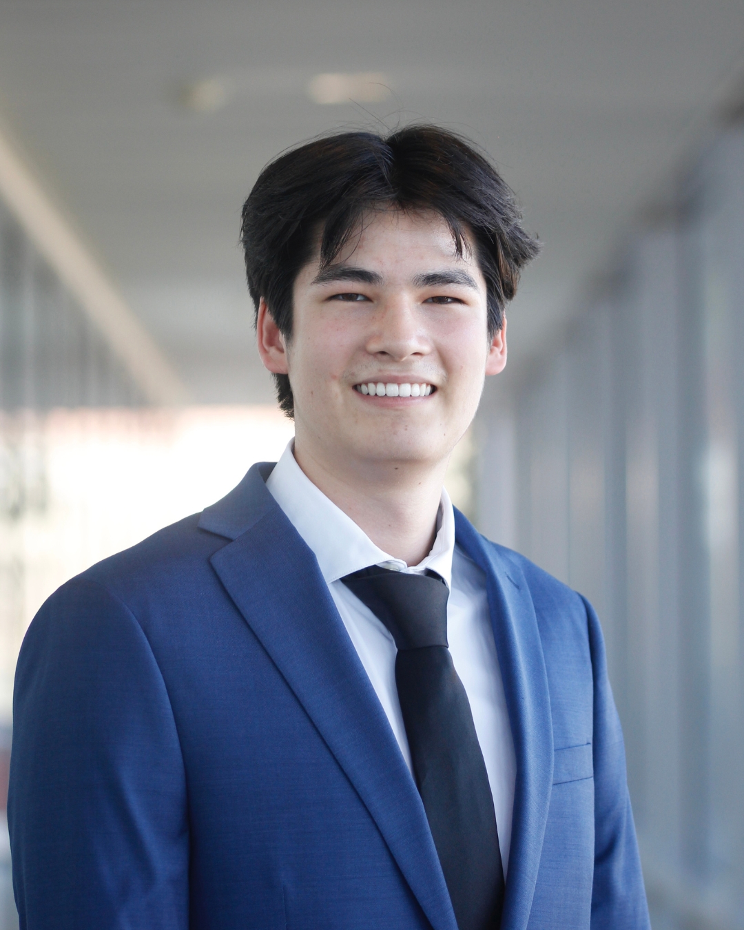 Man with short black hair and blue suit with black tie standing in front of walkway. 