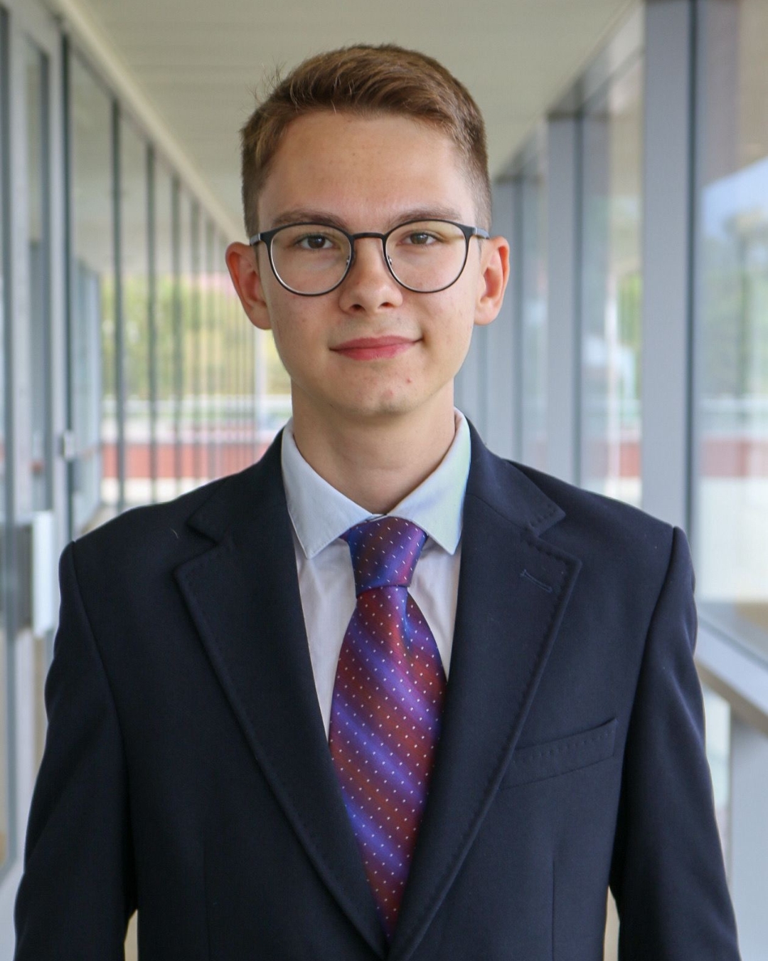 Man in black suit with purple tie wearing glasses and short brown hair standing in front of walkway.