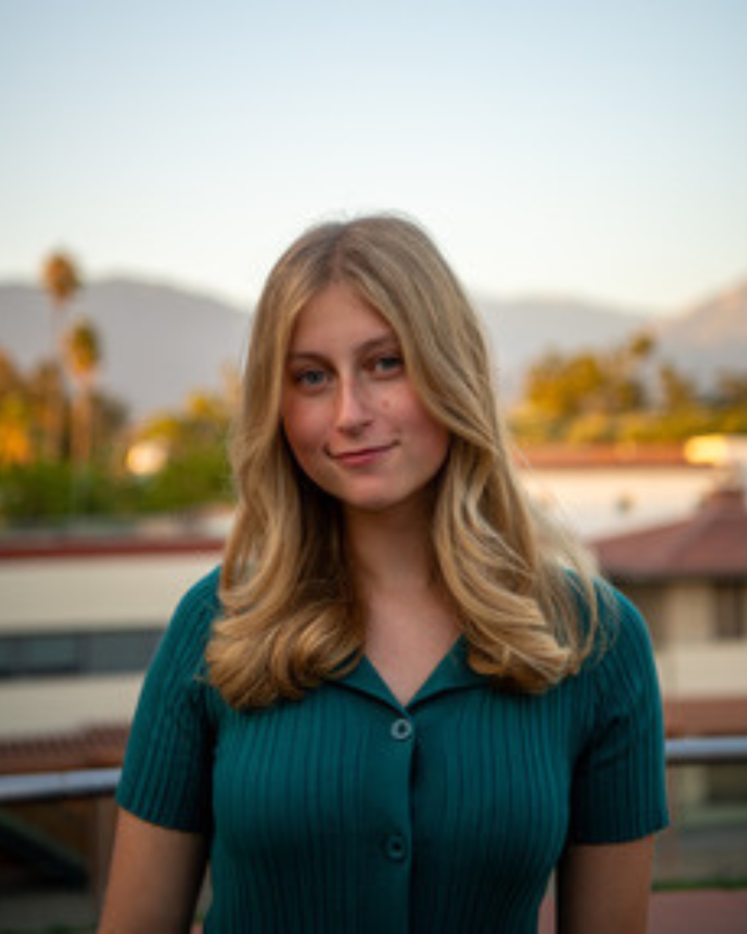 Girl standing in front of rooftop view wearing green top with short blonde curly hair.