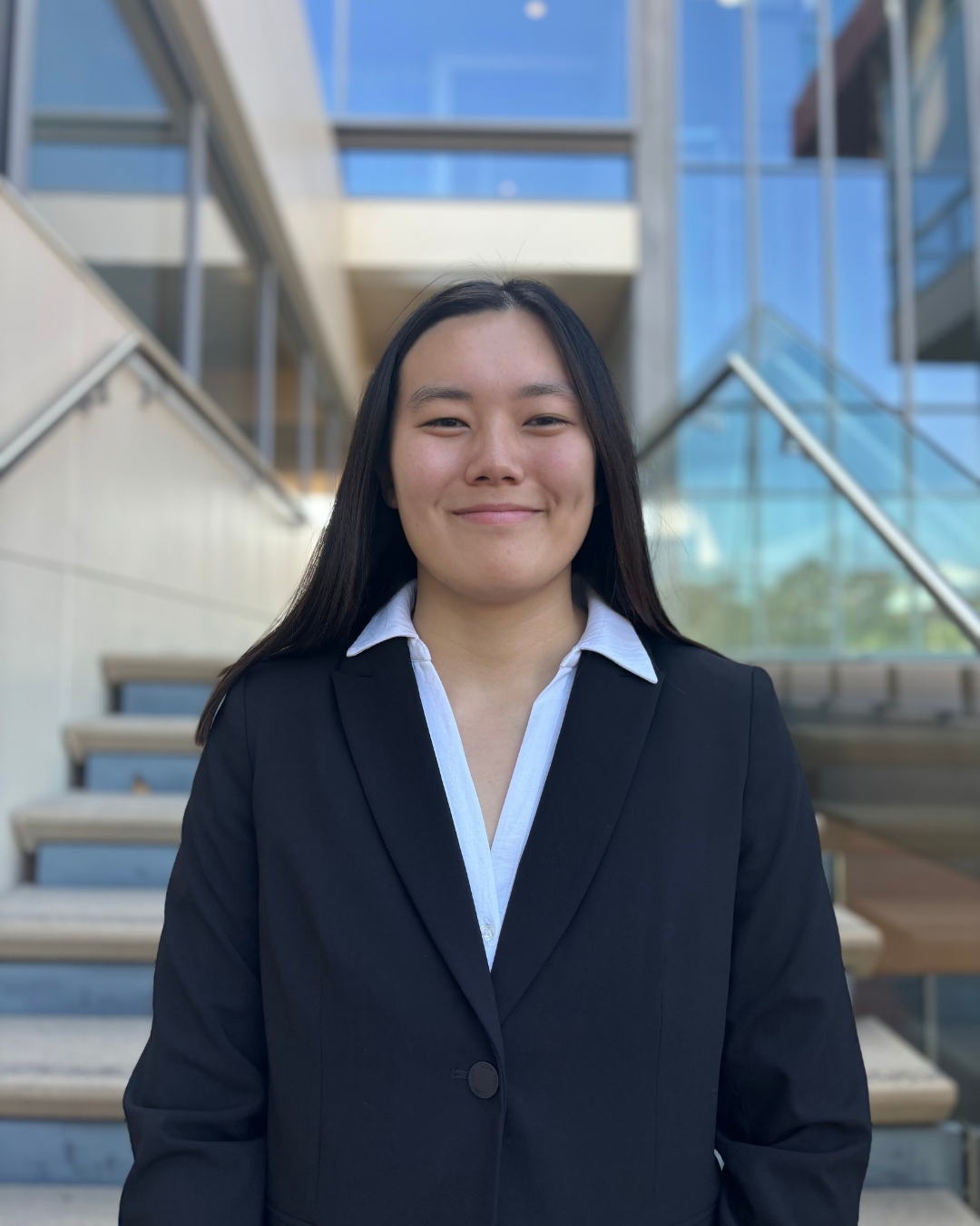 Girl in navy blazer and white top with long black hair standing in front of stairway.