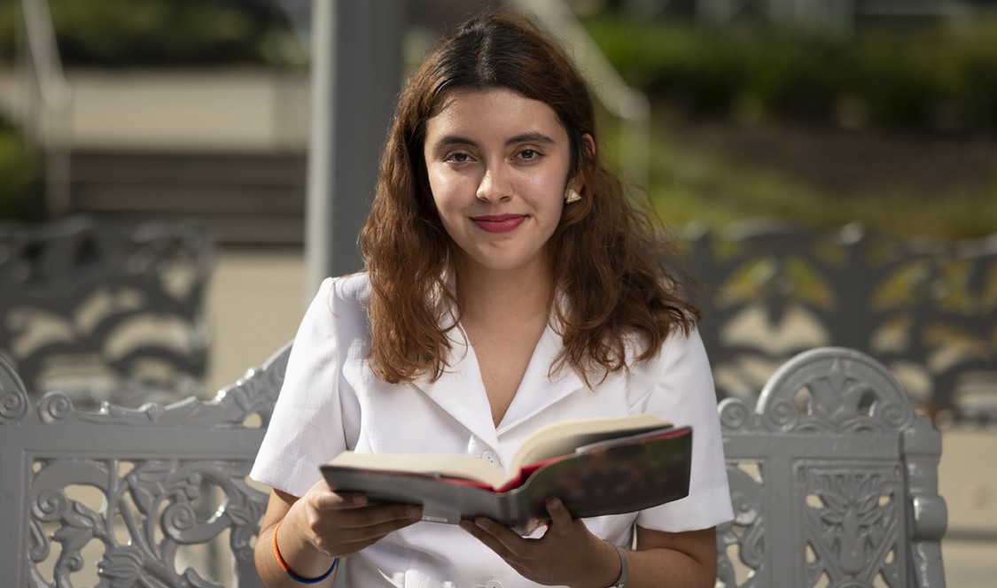 Marycarmen Montarez ’22 holds open a book while sitting on a bench from Artist Chris Burden’s Meet in the Middle public art sculpture.