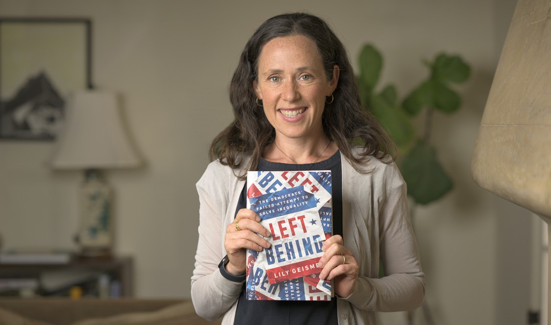 Professor Lily Geismer photographed at home, smiling and holding up her latest book, "Left Behind."