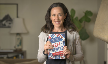 Professor Lily Geismer photographed at home, smiling and holding up her latest book, "Left Behind."